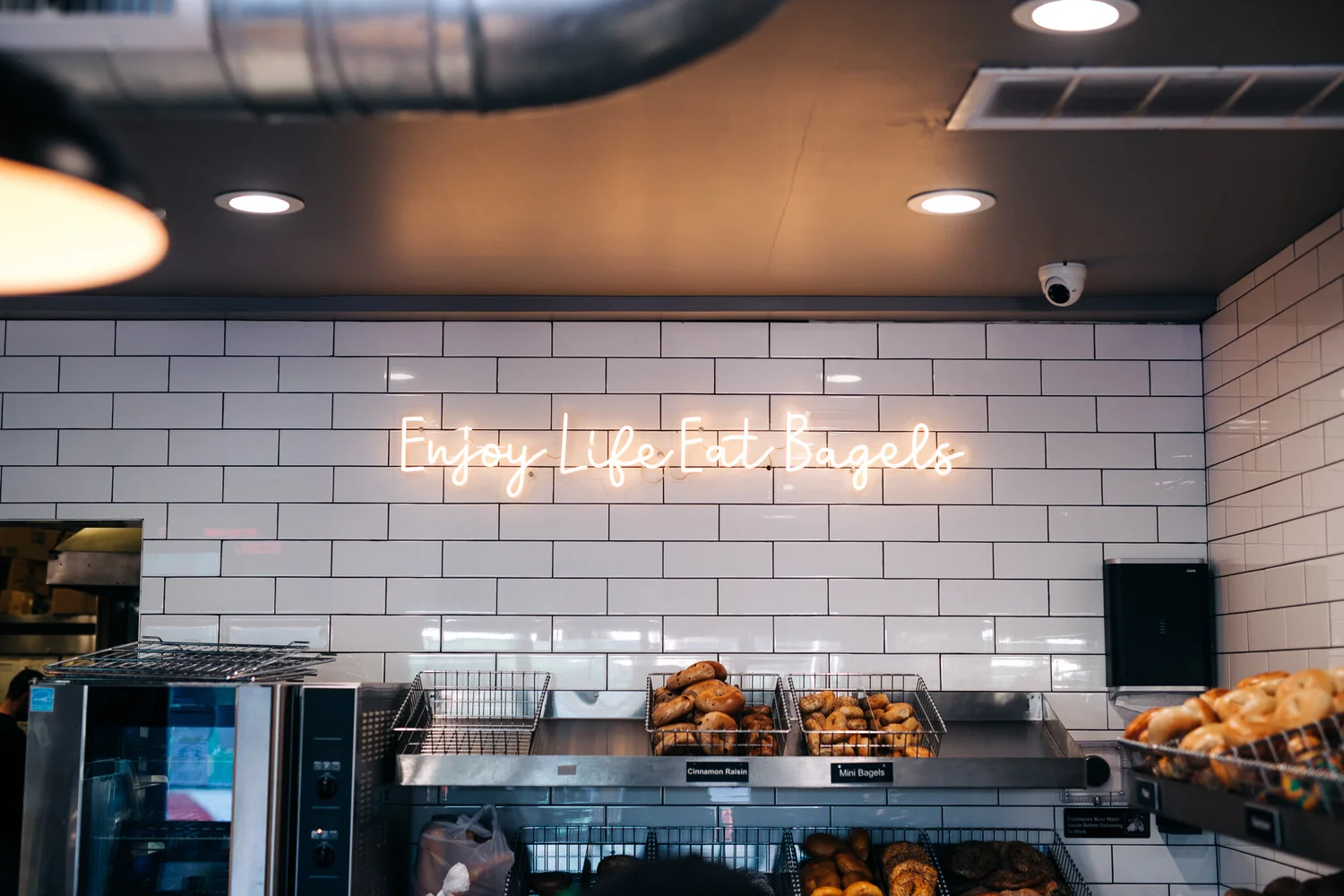 Not Just Bagels shop interior with an Enjoy Life Eat Bagels neon sign over the bagel case